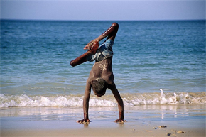 Image of a person with a disability doing a hand-stand on a beach with waves and sand. The image is entitled "Shore of Life" by Franck and won 1st Prize Colour at the WHO Photo Contest: Images of Health and Disability, 2004/2005.