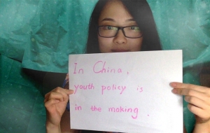 A young woman with glasses holds up a sign that says in pink, "In China, youth policy is in the making"