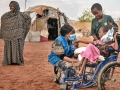 8岁的小姑娘伊克兰坐在轮椅上，朝世界粮食计划署瓦吉尔外地办事处主任勒奈特·瓦蒂蒂（Lynette Watiti）微笑，她的母亲哈莉玛（Halima）在一旁看着他们。