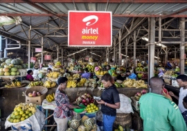 An advertisement for a mobile phone money transfer company hangs over stalls of fruit and vegetables at a covered market in Kigali, Rwanda. Photo: Panos/Sven Torfinn