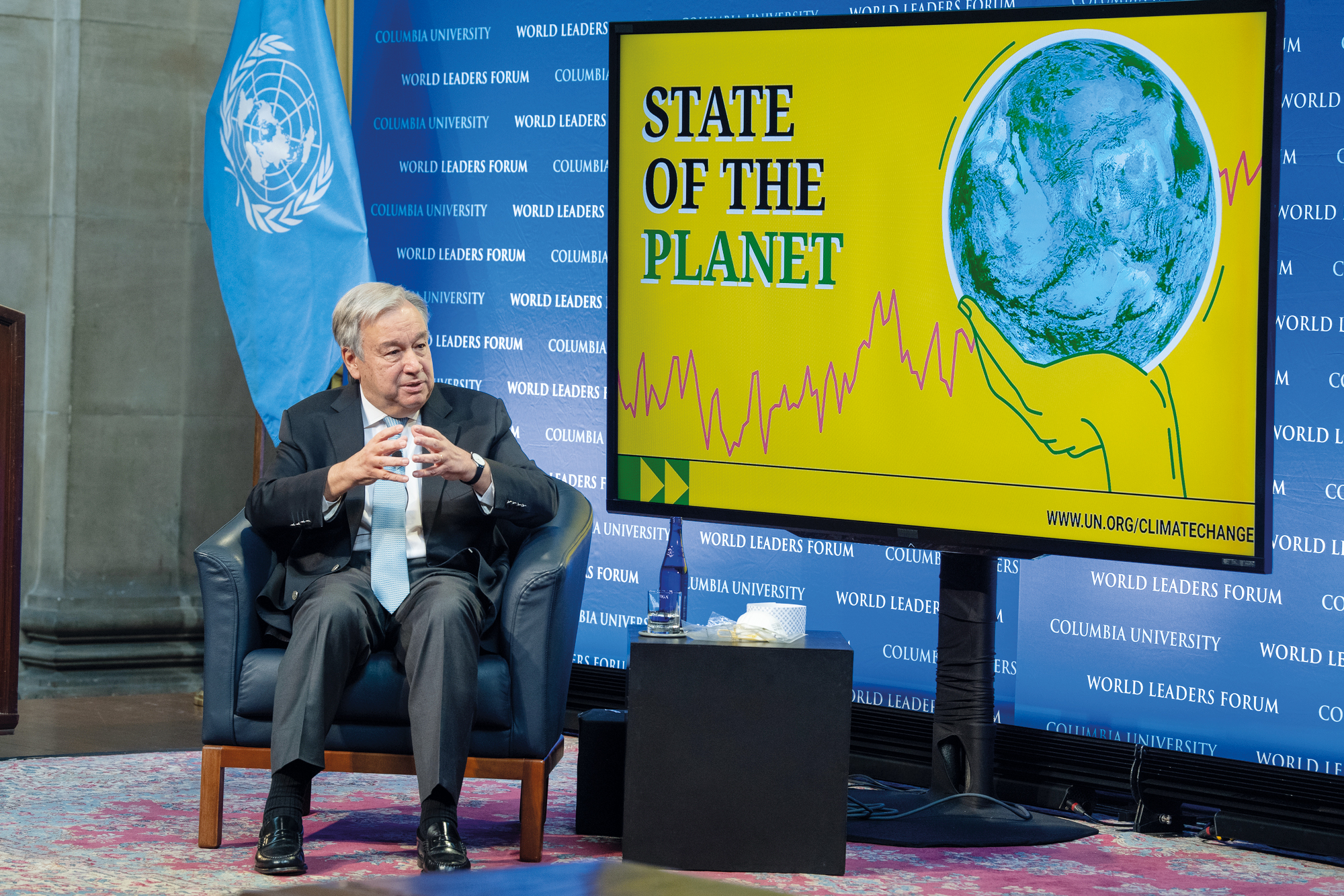 The Secretary-General sits and speaks to someone out of frame. Behind him, a TV screen displays the words "State of the Planet" and a picture of the globe.