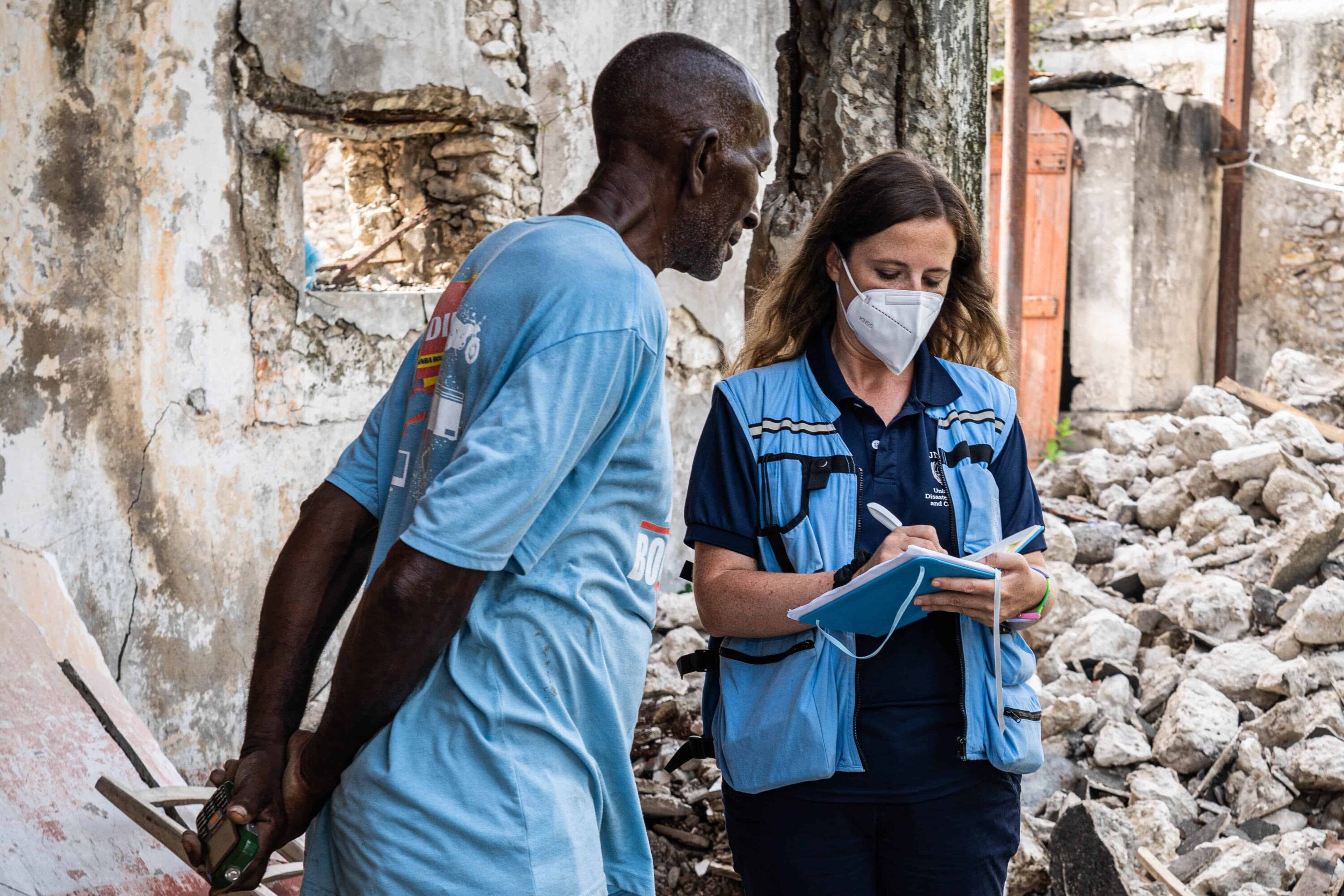 A man and an aid worker stand facing each other. The aid worker is writing in a notebook. Behind them are ruined buildings and rubble.