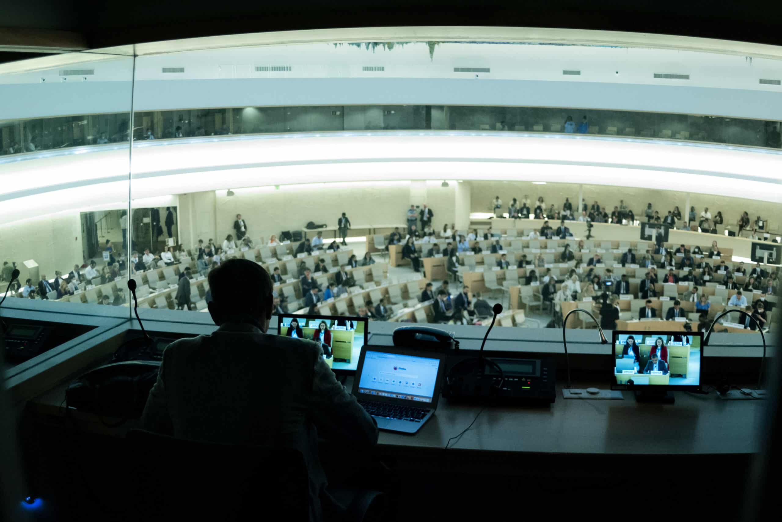 A person sits with his back towards the camera in a booth with a large window overlooking a conference room. The person is seated by a desk with two laptops and microphones.