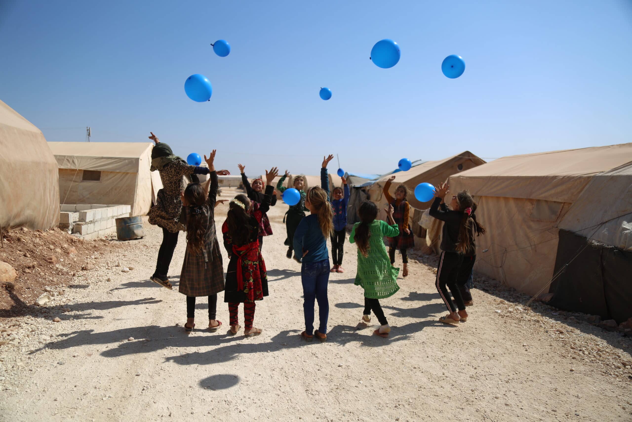 A group of children form a circle outside. They have their hands raised and are jumping up towards several blue balloons floating above them.