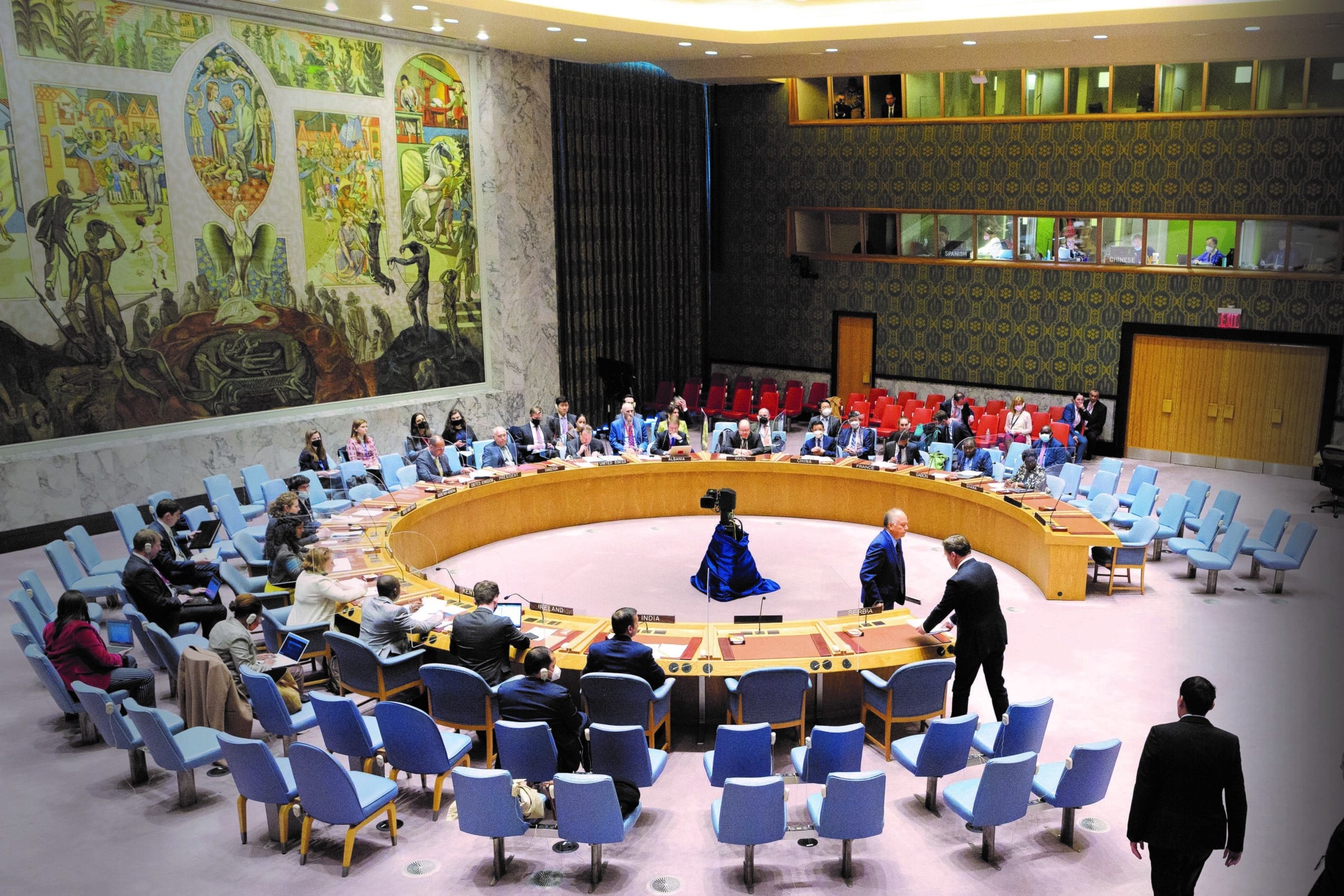 A wide view of a conference room, with tables in a circle. Several people are moving around the room. On the left wall is a large tapestry.