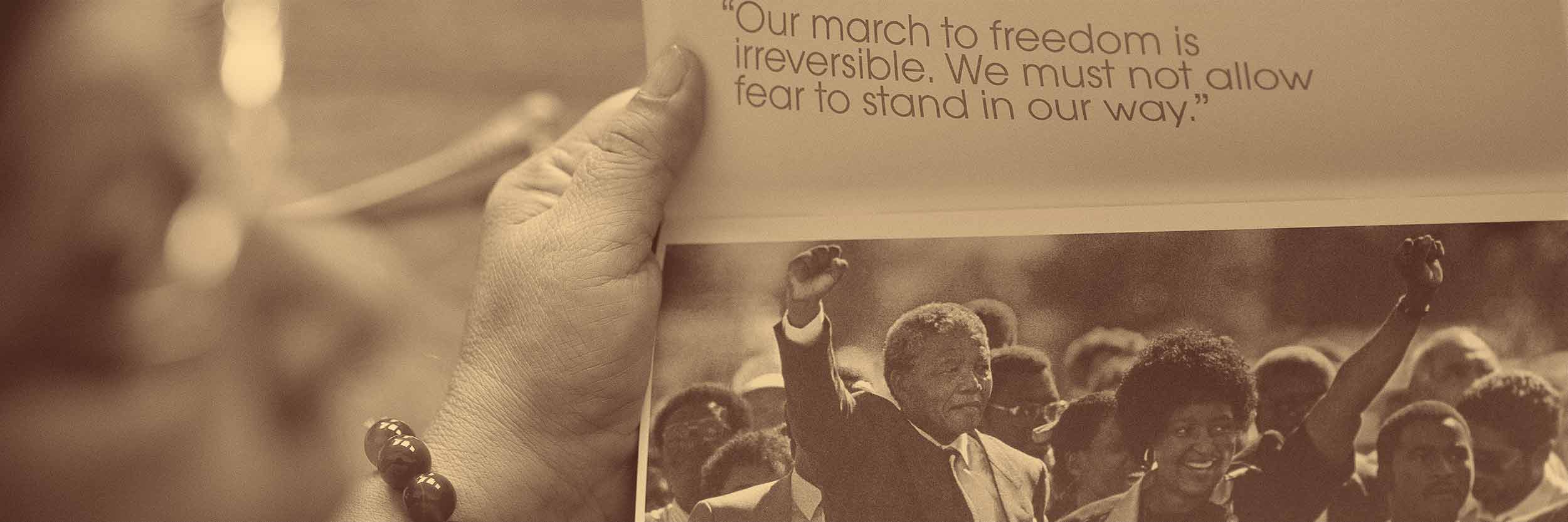 A woman holds up a booklet with a photo of Nelson Mandela hoding up his fist and his wife's had, while surrounded by people.