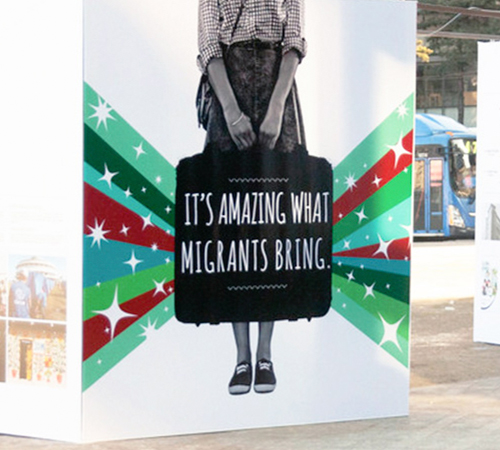 Exhibición pública de grandes paneles cuadrados con ilustraciones de individuos llevando una maleta con la inscripción 'es increíble lo que aportan los migrantes'