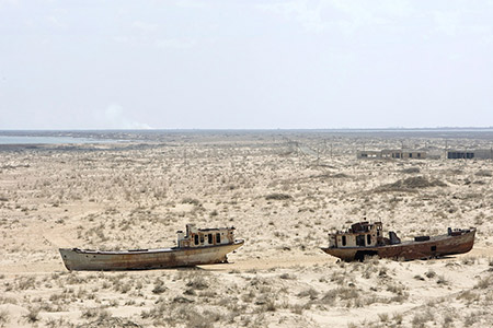 Two ships lie stranded in a vast field. There are small houses and powerlines in the background. 