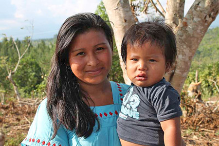 A young woman in a blue dress stands in a coffee plantation with her infant child who is dressed in a grey shirt. There is a large tree behind them. 