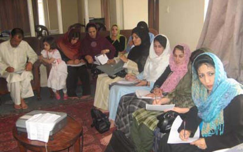 A group of older woman sit on two lounges. All women are holding pieces of paper and are writing notes with pencils. There is a table in the middle of the room with a projector.
