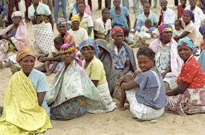 A large group of women are sitting on the ground, which is covered in sand. Some of the women are breastfeeding their children.