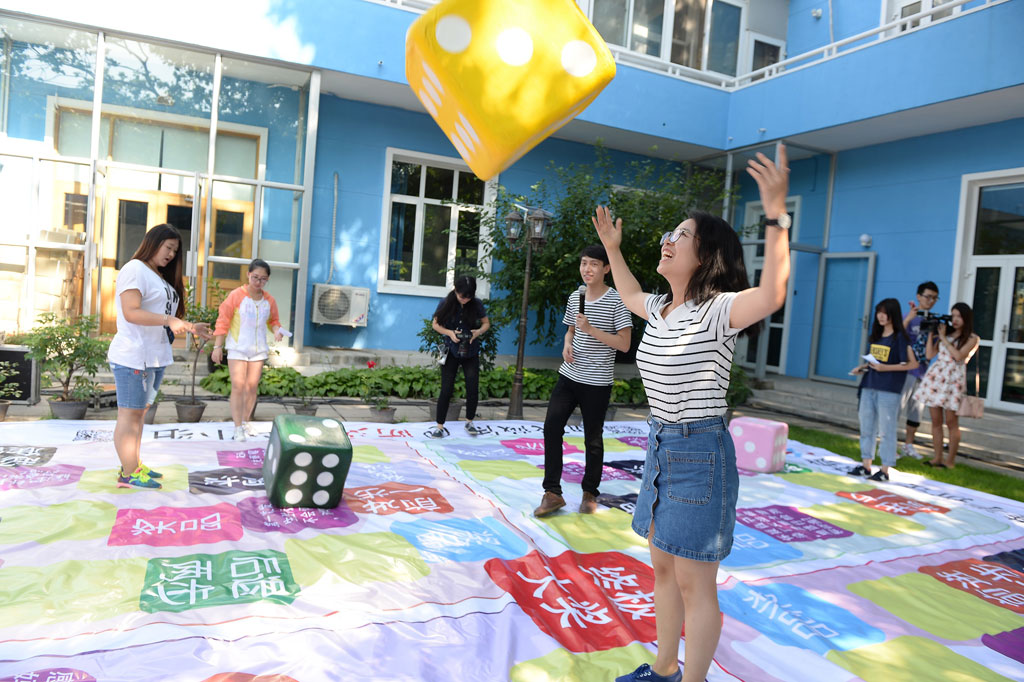 A young woman throws a large yellow dice in the air. There is a young man standing behind her with a microphone. Several other young people are standing in the open air space, surrounded by a blue building.