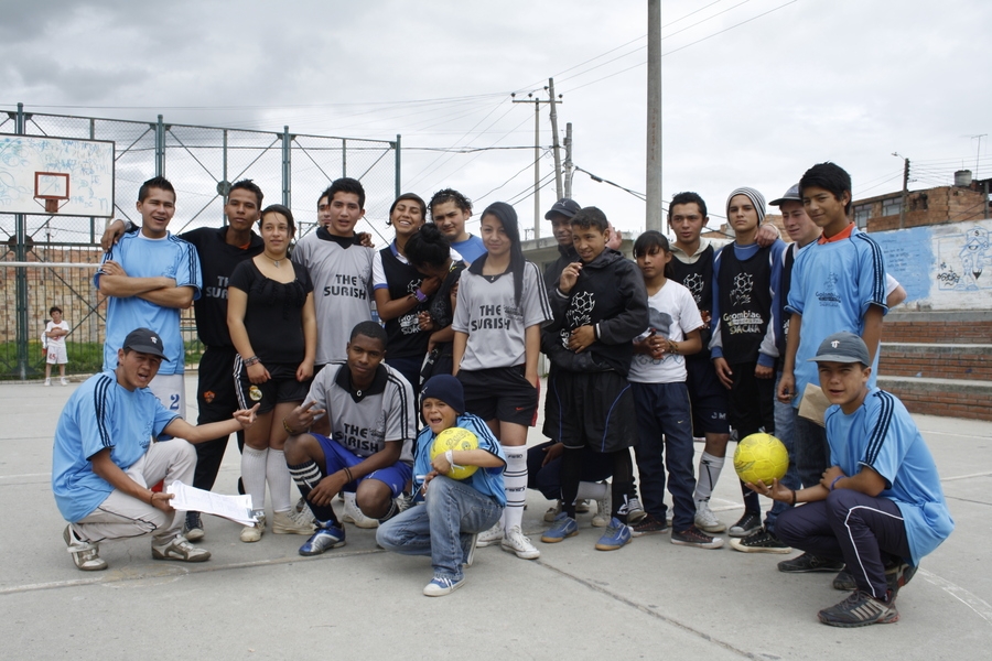 A group of adolescents dressed in blue and grey uniforms stand in a basketball court surrounded by brick walls. Two of the boys sitting at the front of the photo are holding bright yellow soccer balls. The sky above them is dark and grey.