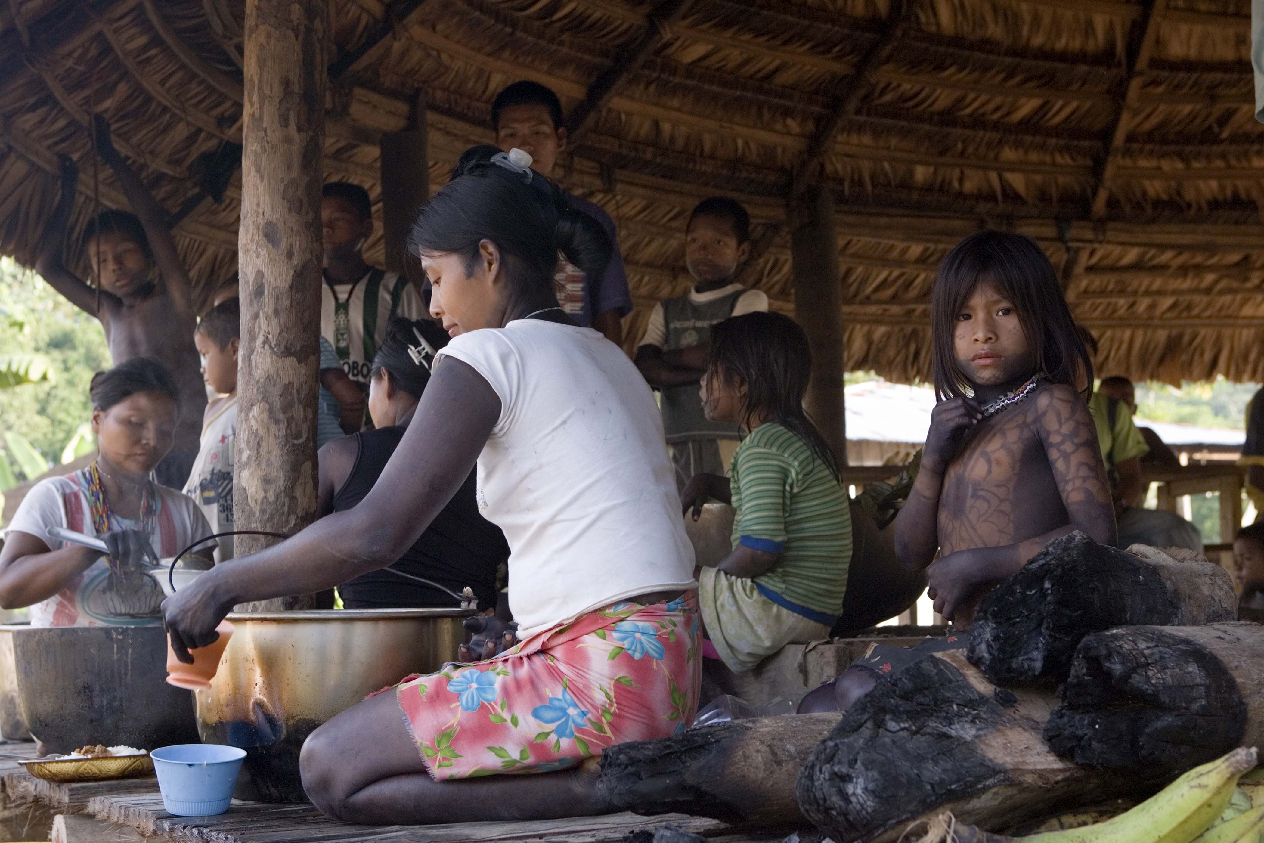 A woman cooks in a pot surrounded by young children who are watching her cook. They are all sitting in a hut.