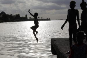 A boy is smiling as he jumps into the water, while several boys are cheering him on.