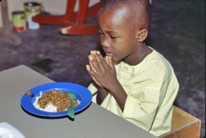 A boy offers a thanksgiving prayer before eating his meal at the refugee camp in Buduburam, Ghana. A bowl has been placed in front of him, filled with rice and meat.