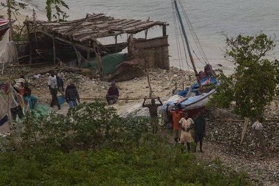 A group of people wave to the camera from a distance, surrounded by damaged buildings and boats which were affected by the cyclone. They are adjacent to the ocean.
