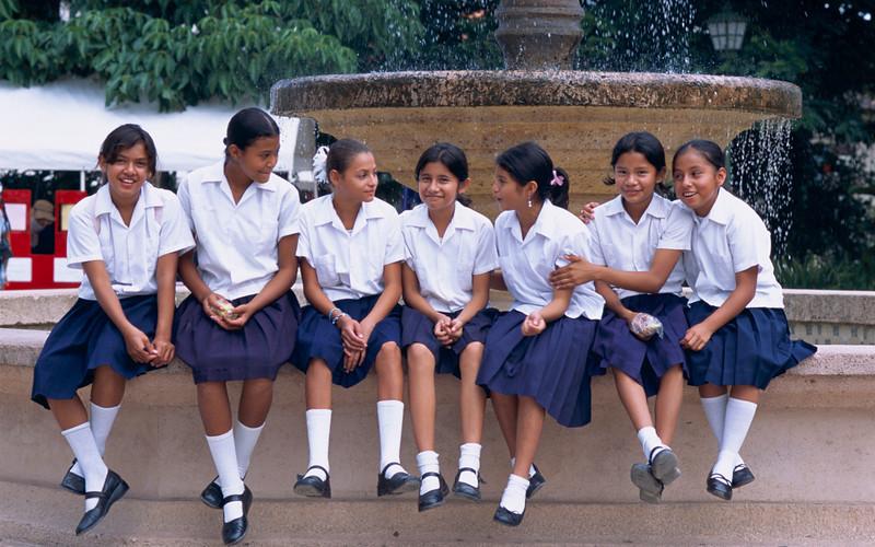 Seven young girls dressed in a school uniform sit on the edge of a fountain while chatting to each other.