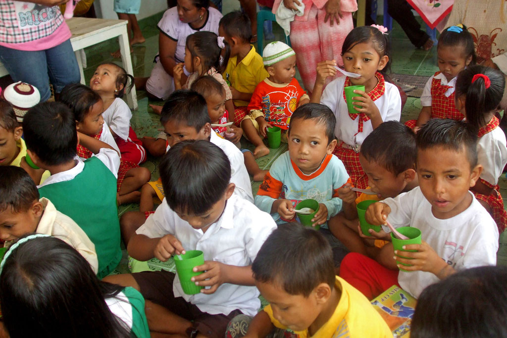 Young children sit on the floor of a classroom eating a meal out of small green cups with a spoon.