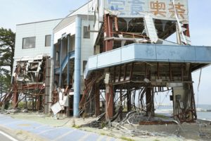 A house stands on the edge of the land next to a bay of water. It is damaged from a tsunami, with wires and bricks laying underneath it.