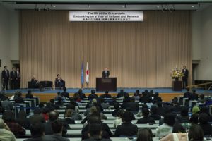 A man on a large blue stage stands in front of an audience. Behind him is a UN flag and a Japanese flag.