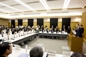 A speaker at a podium holds a microphone as he addresses a group of students and delegates sitting at desks surrounding the podium. They are seated in a large room with dark black curtains.