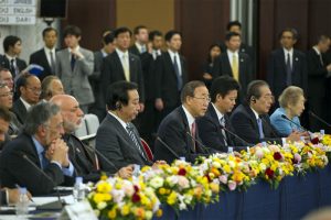 Seven panellists sit at a table facing microphones. There are bouquets of flowers lining the edge of the table. Behind the panellists stand a group of men wearing suits.