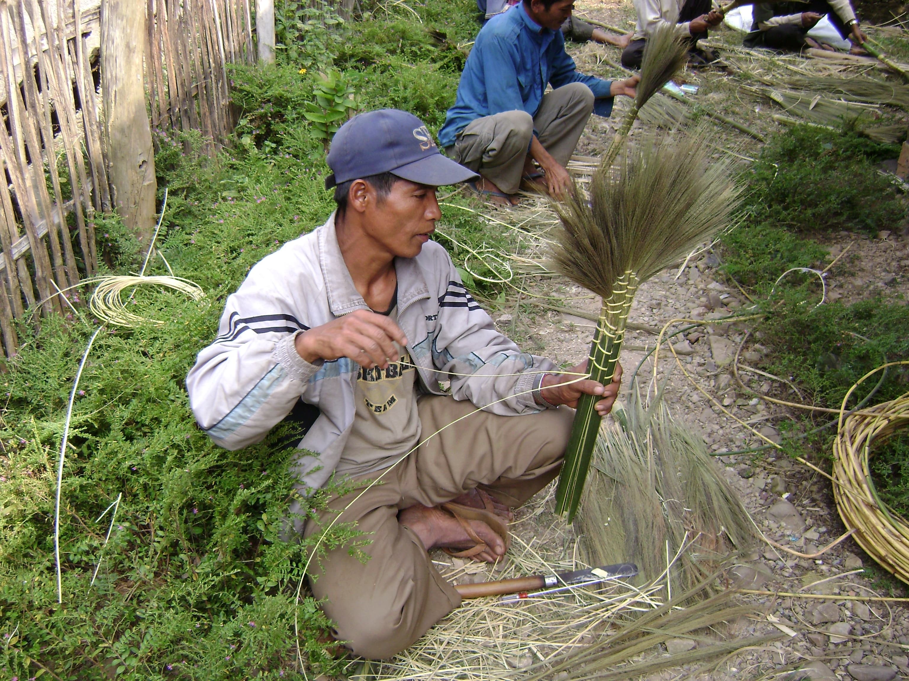 A man sits on the ground with open shoes and is weaving plants using bamboo and string. There are several other men behind him who are also weaving.