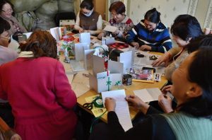 Older women sit around a table engaging in an art and craft activity. The table is full of paper and cards and the women are cutting out shapes and painting.