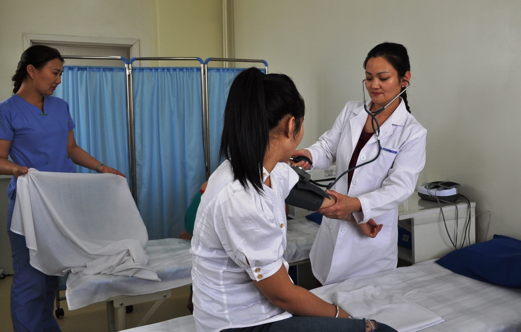 A female doctor wearing a white lab coat with a stethoscope checks the blood pressure of a young Mongolian woman sitting on a bed. There is a nurse behind them wearing a white uniform and she is folding a sheet.