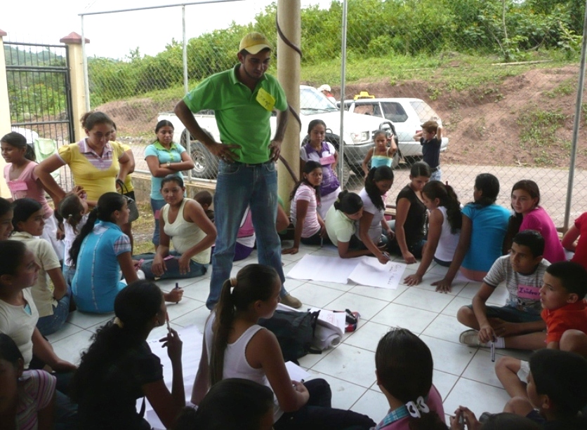 A man stands with his hands on his hips in front of a group of children sitting around him in a circle. Some of the children are drawing on large white sheets of paper.