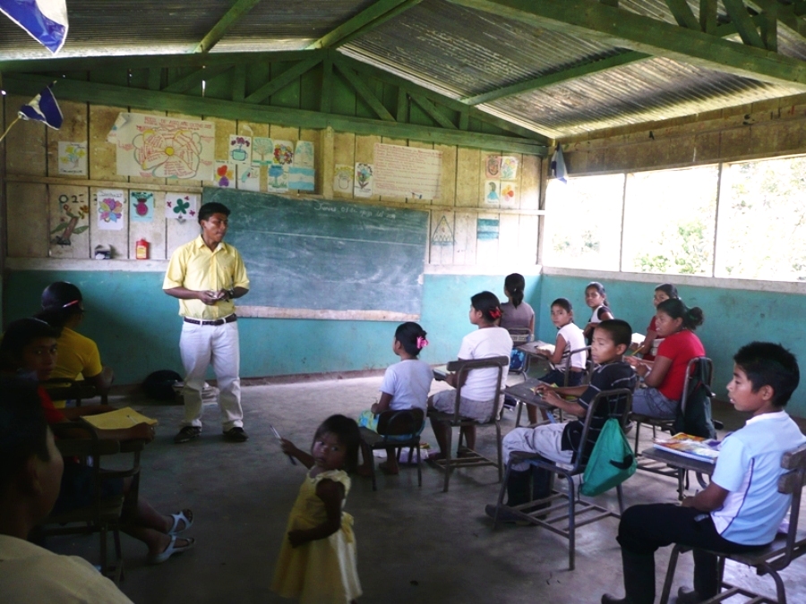 A man teaches a class of young students. He is standing in front of a chalkboard, with the students seated at small wooden desks in front of him.