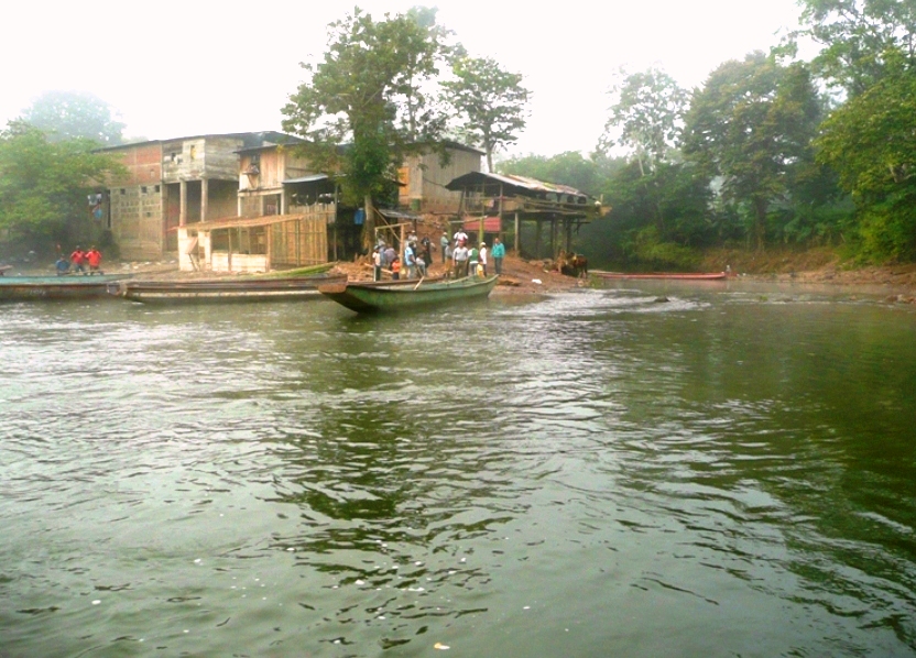 A river wraps around a group informal settlements. There is a canoe floating in the river, with several individuals watching on.