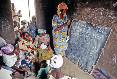 Women are pictured attending an adult literacy class in a village near Bauchi. A woman is writing on a chalkboard which is leaning against a wall.
