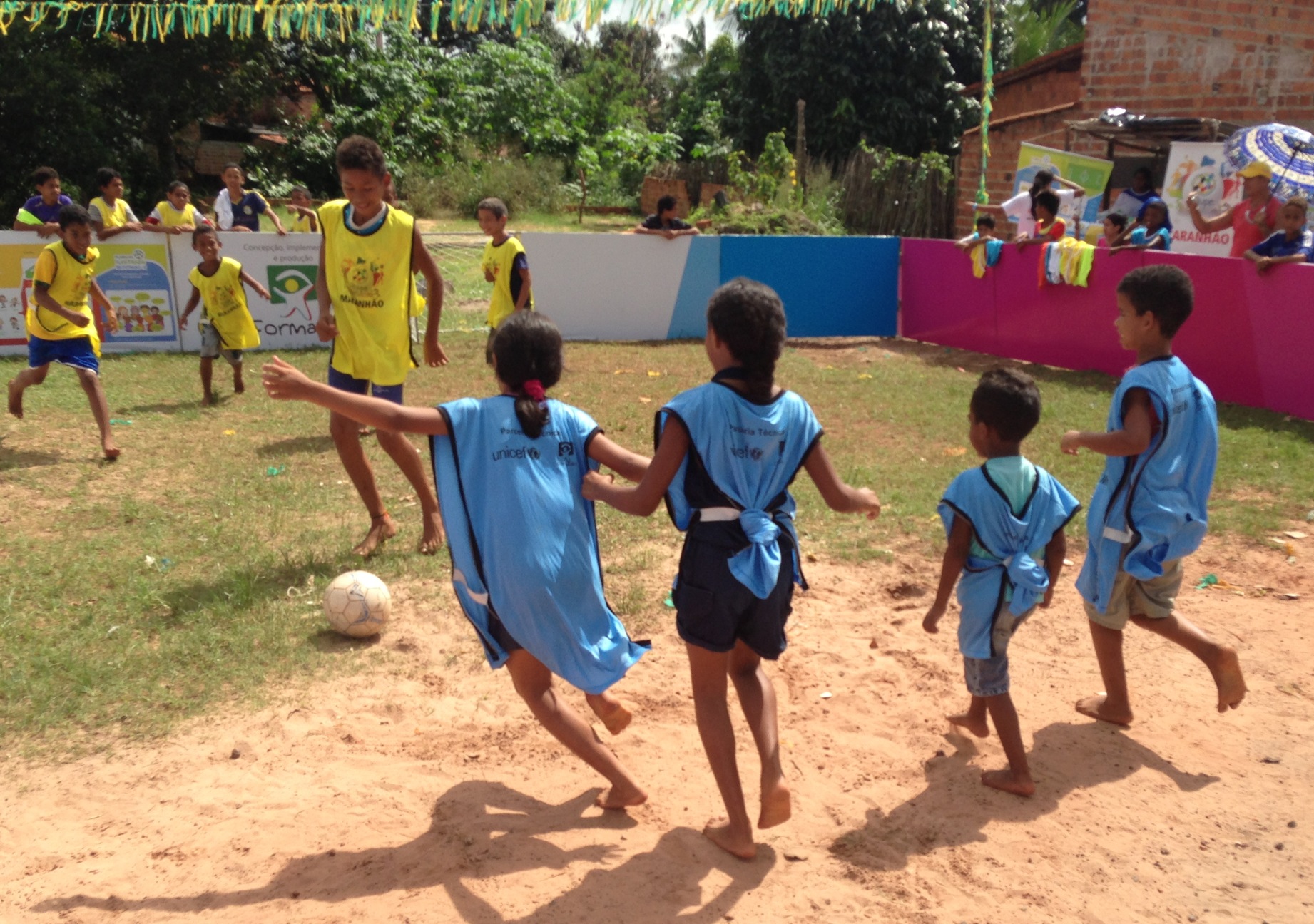 Three young girls dressed in blue uniforms play soccer against a team of young boys dressed in yellow. A young girl is running across the field to kick the soccer ball. There are several children watching the game from behind a fence.