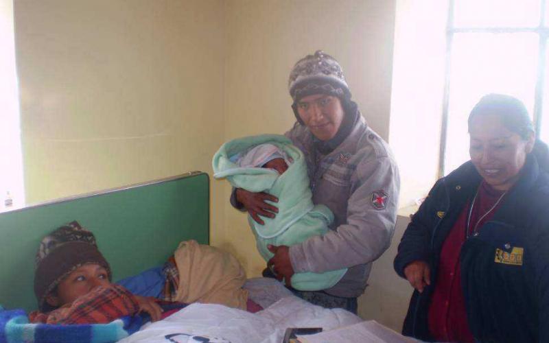 A smiling male doctor, wearing a stethoscope and blue jacket, tends to a new born baby crying on a bed.