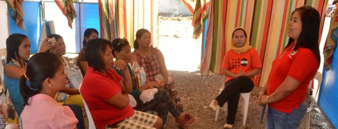 A woman in a red shirt stands before a group of young women who are sitting in white plastic chairs. They are in a room surrounded by colourful sheets of fabric.