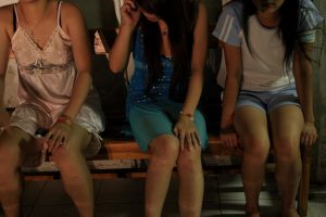 Three girls sit on a wooden chair. One girl has her hand resting on her leg and a phone to her ear. Their faces are obscured.