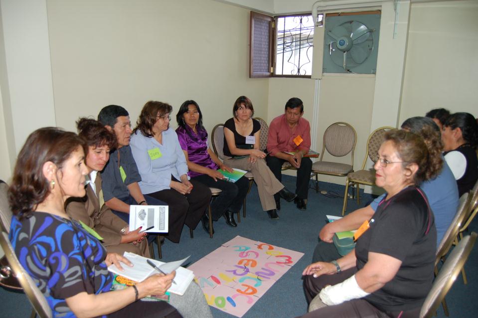 A group of older men and women sit in chairs in a circle formation. They are holding colourful classroom resources. There is a handmade poster sitting on the floor in the middle of the circle.