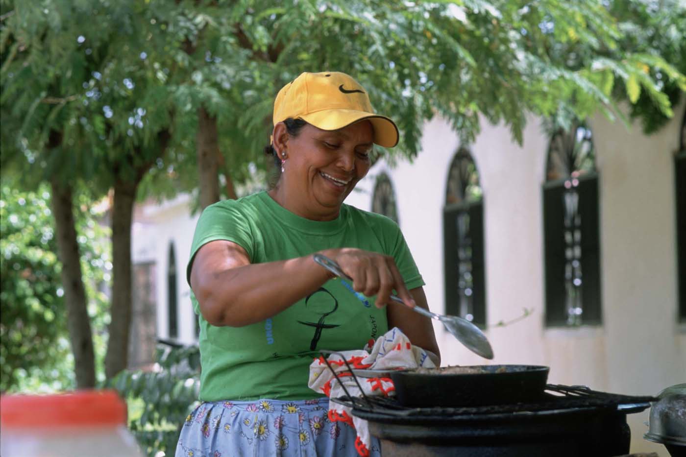 A woman dressed in a green shirt and yellow smiles as she cooks a meal in a saucepan.