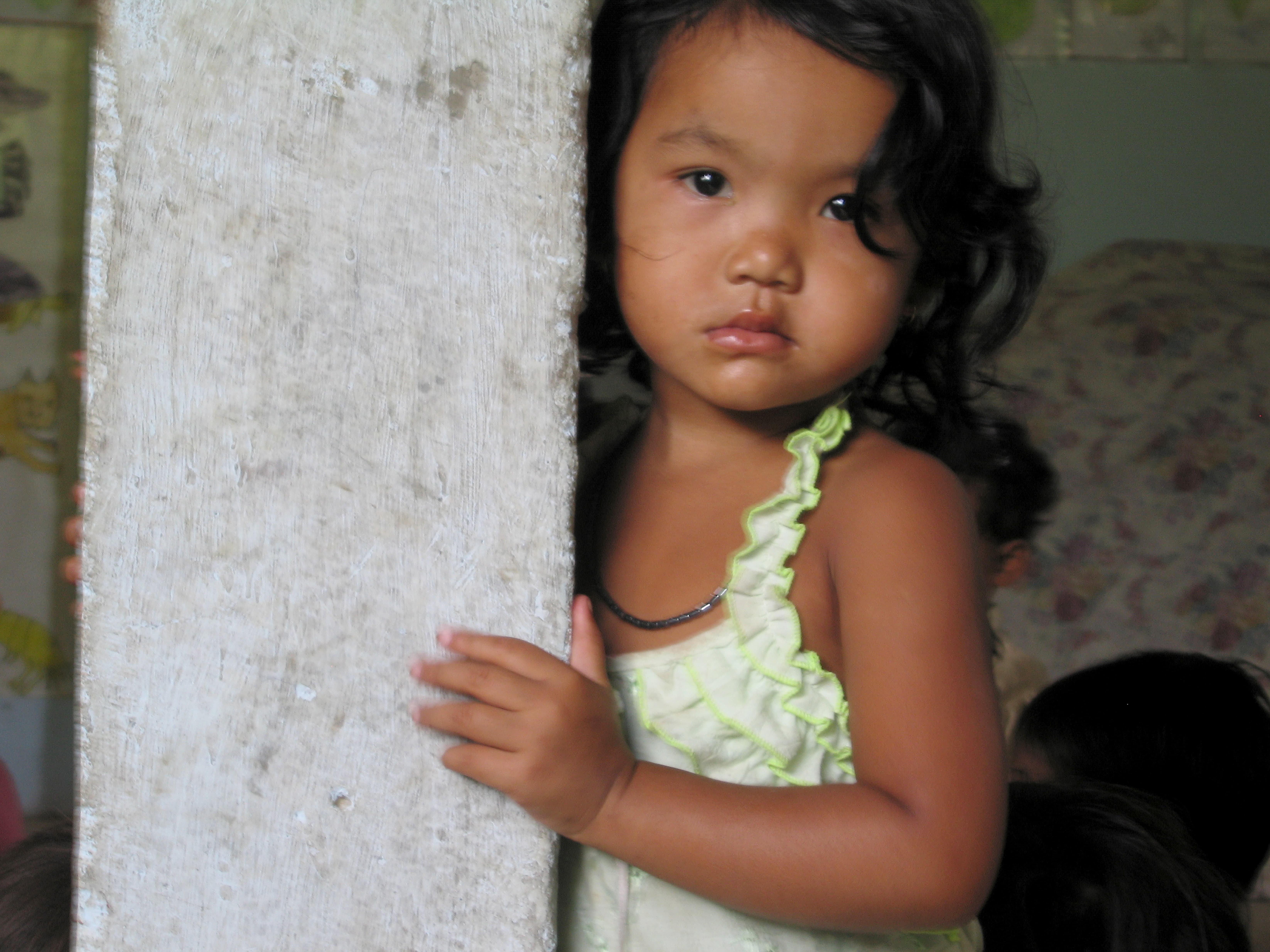 A young girl stares at the camera as she holds onto a wooden pole. She is wearing a green and white singlet and a black necklace.