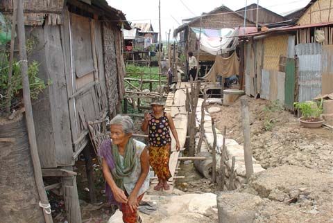 Two elderly women walk through an undeveloped village. They are surrounded by dirt. There are clothes hanging on string in the background.