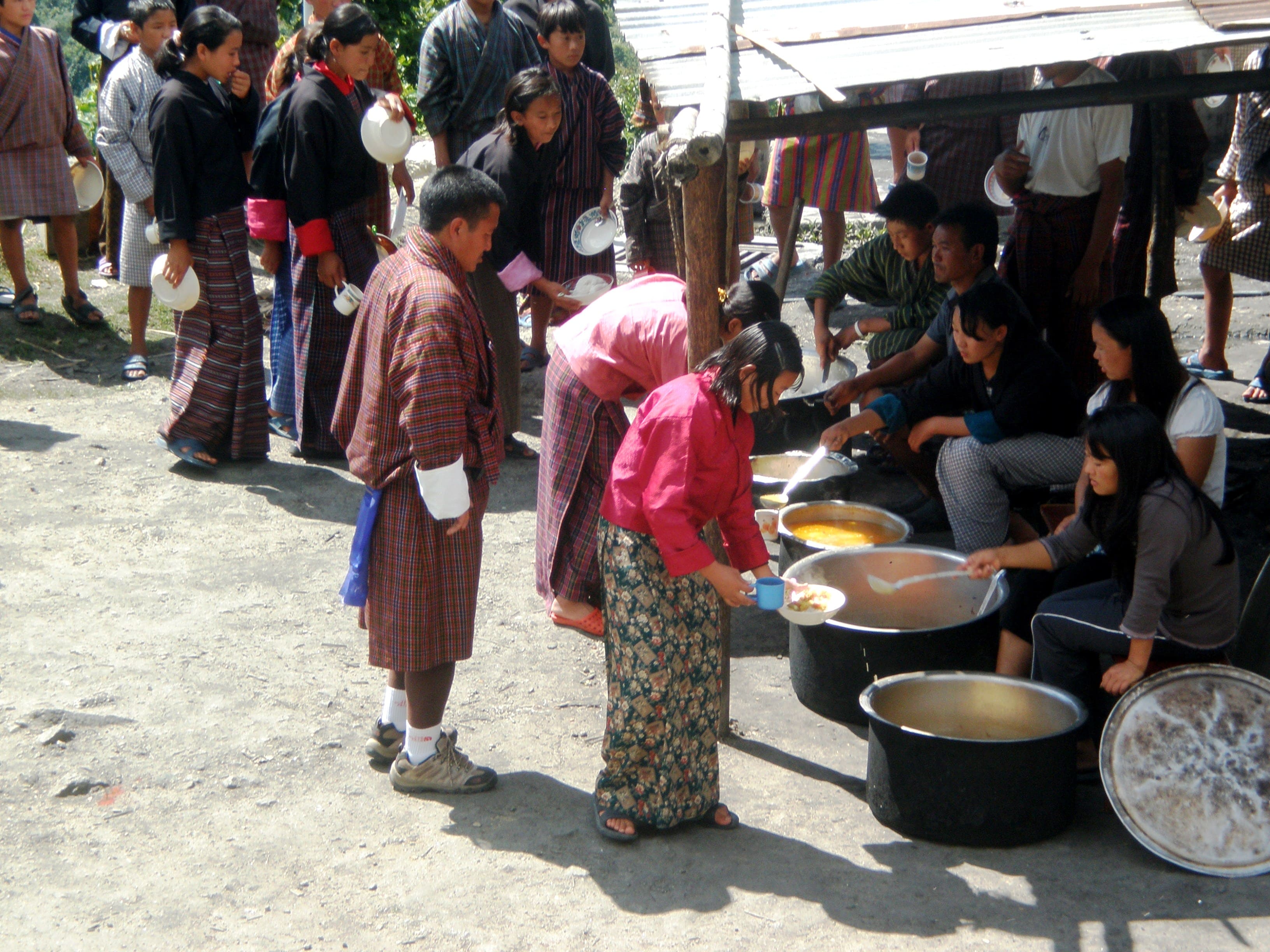 Several men and women hold bowls and walk up to a food serving station. There are girls sitting next to large bowls of food at the food serving station and are serving the food with large ladles.