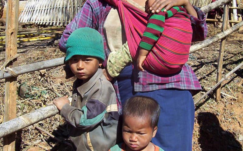 A woman leans against a wooden fence and is carrying a baby in a sling. Two younger children are standing in front of her. There is a paddock and a hut behind the wooden fence.