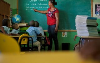 A teacher stands in front of a green chalkboard and points to text that has been written on the chalkboard. Four young students on her right are sitting at their desks and are reading from the chalkboard.