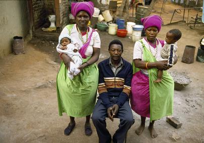 A man sits on a chair surrounded by two older women who are carrying children.