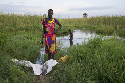 A young woman shows off a fish she just caught in a lake. Behind her, a child is wading through the lake and looking for fish. The lake is surrounded by grass.