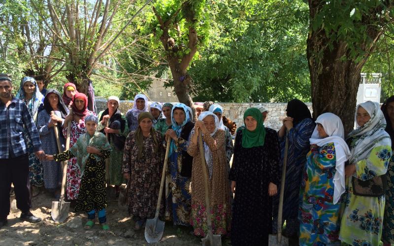 A dozen women stand with shovels in a field. There are large trees lining a stone wall.