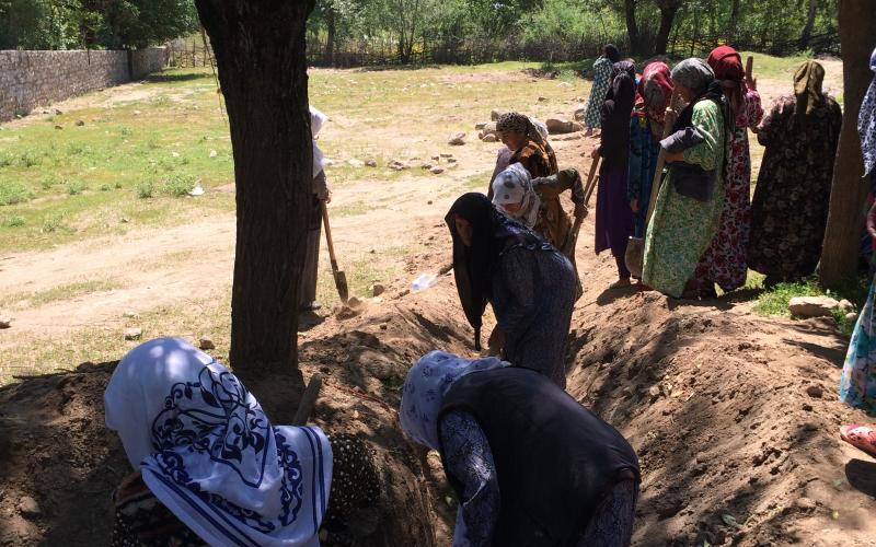 A group of women dig into a dirt mound surrounding a large tree. There are several women watching from the side. They are standing in a large field surrounded by wooden and brick fences.