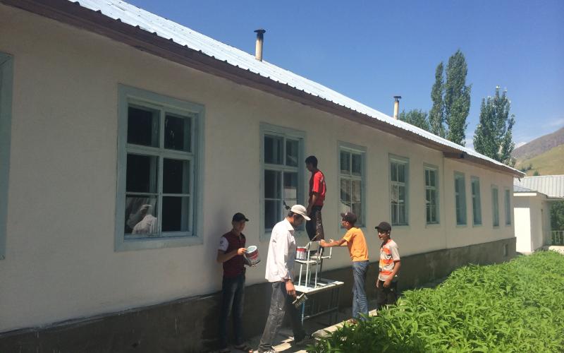 A group of men stand next to a long white building with an aluminium roof. One of the men is standing on a table which he is using as a ladder to paint the wall.